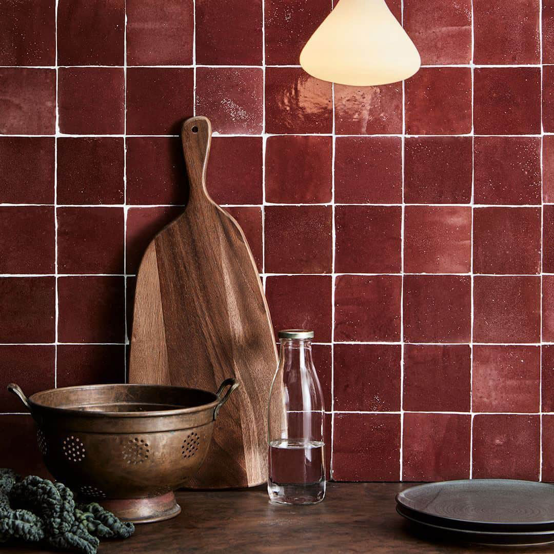 Kitchen scene with a wooden cutting board, colander, glass bottle, and greenery against a red tiled wall.